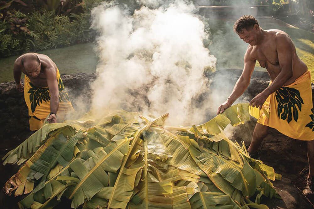 2 men arrange leaves atop traditional imu.